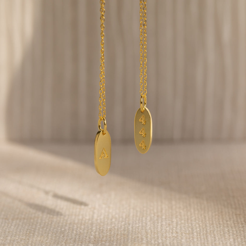 Two Old English Oval Tag Necklaces in gold, one engraved with the initial "A" and the other with "444," are displayed against a neutral background.
