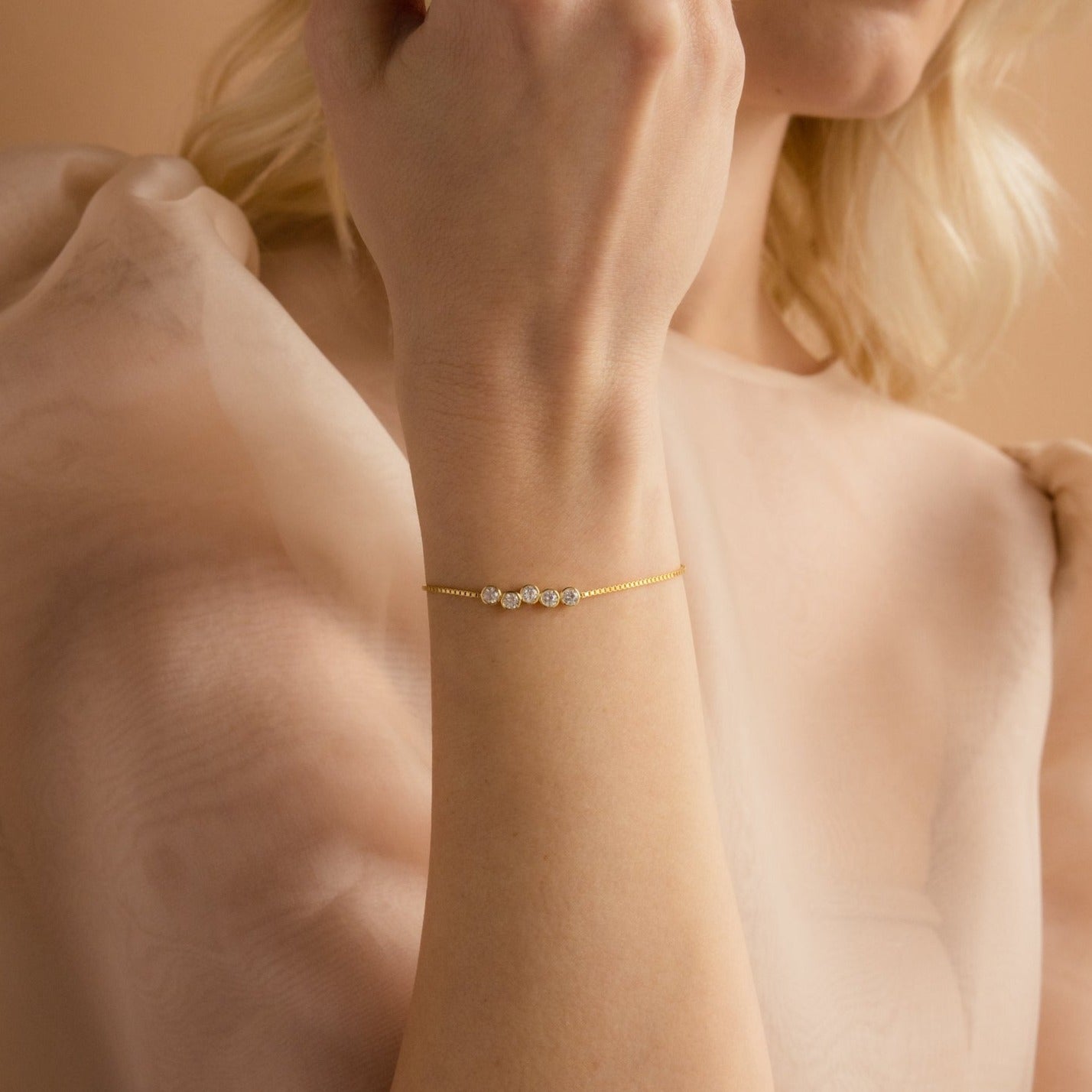 A woman wears the Cluster Birthstone Bracelet, a delicate gold piece featuring three gemstones, styled with a sheer, light-colored blouse.