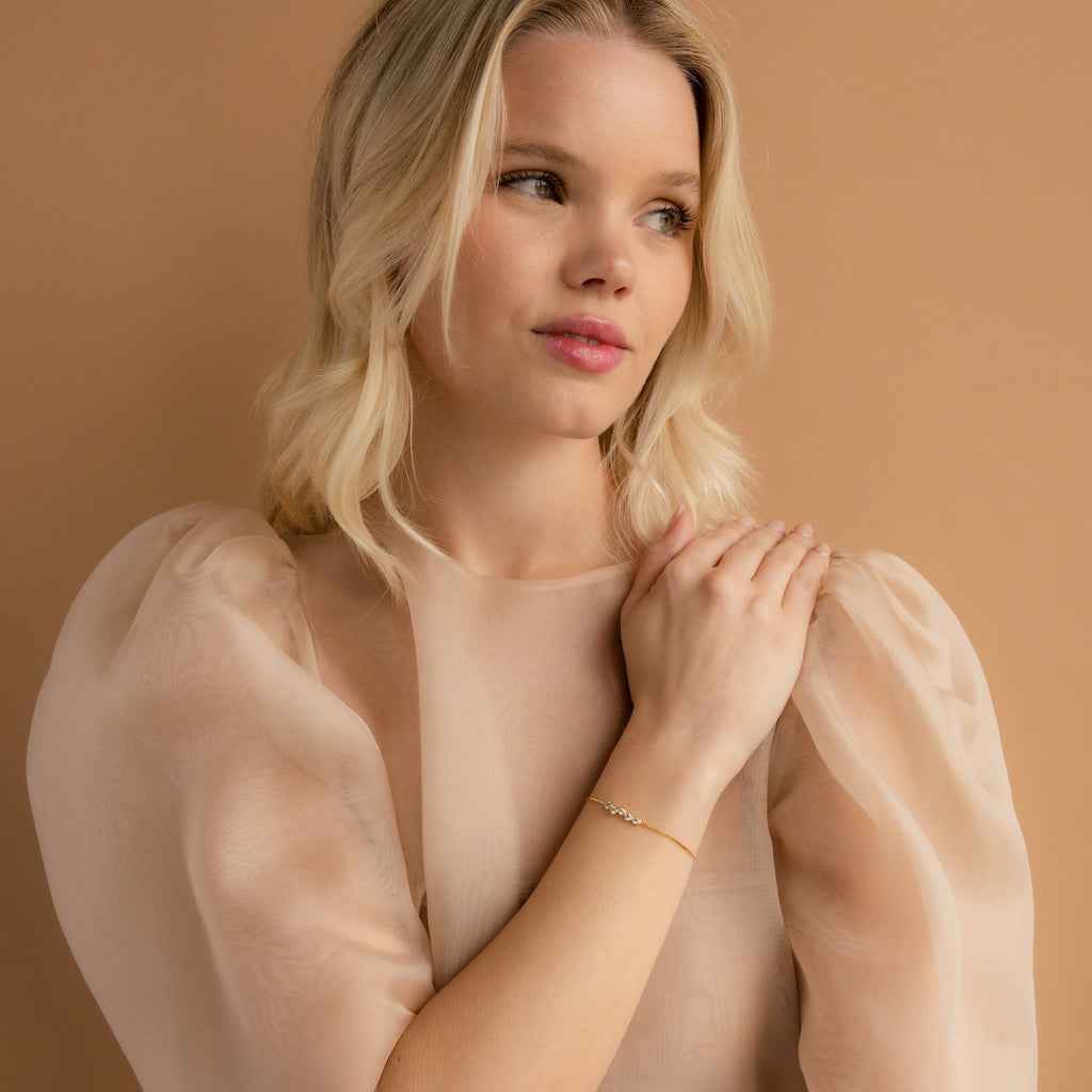 A blonde woman in a sheer, puffy-sleeved blouse poses against a beige background, hands clasped to showcase the Cluster Birthstone Bracelet on her wrist.