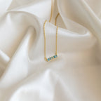 The Initial Morse Birthstone Necklace, featuring blue gemstones, rests on soft cream satin fabric, offering a unique touch of personalized jewelry.