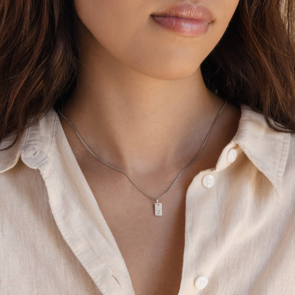 A woman wears a beige shirt and the Old English Mini Tag Necklace, featuring a silver engraved initial on a small rectangular pendant.
