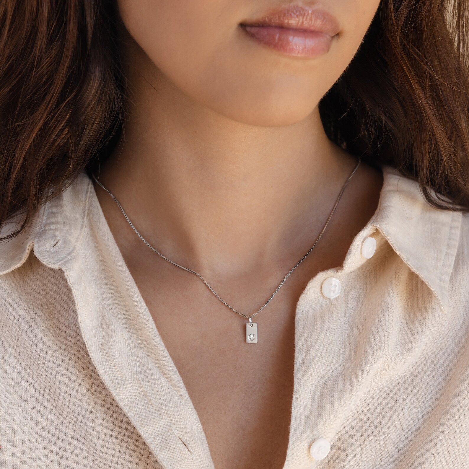 A woman wears a beige shirt and the Old English Mini Tag Necklace, featuring a silver engraved initial on a small rectangular pendant.