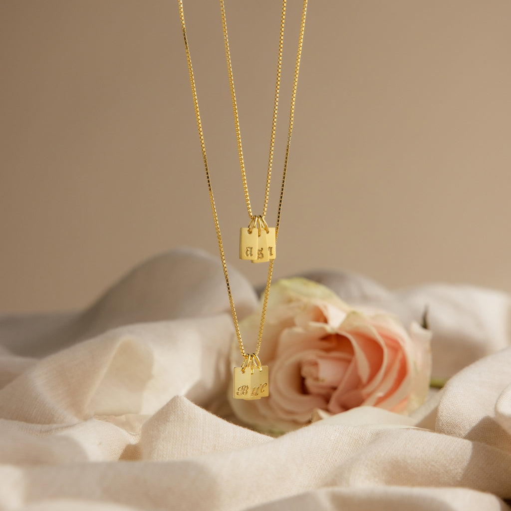 Two Old English Mini Tag Necklaces with engraved initials rest on soft white fabric beside a pink rose, highlighting the elegance of personalized jewelry.