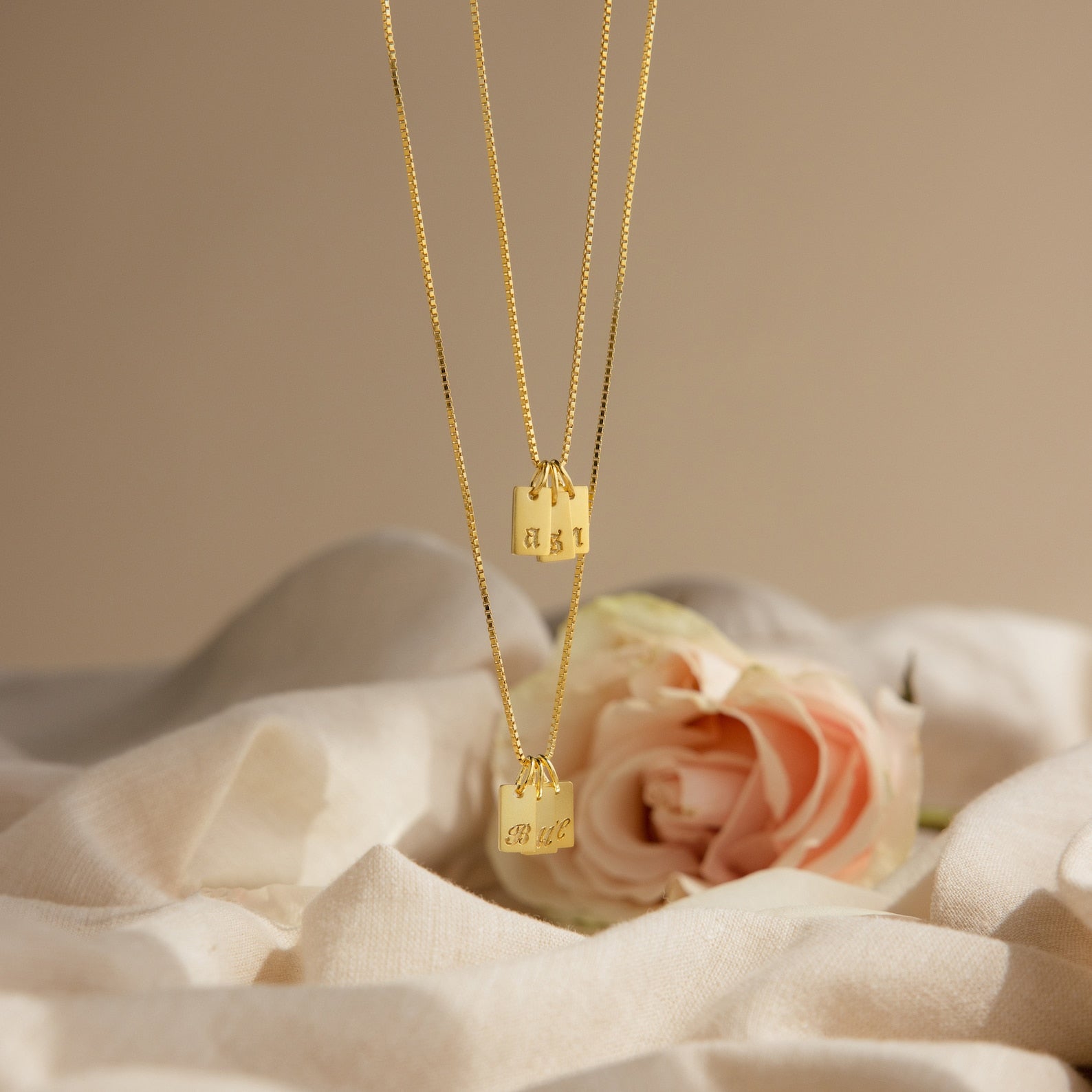 Two Old English Mini Tag Necklaces with engraved initials rest on soft white fabric beside a pink rose, highlighting the elegance of personalized jewelry.