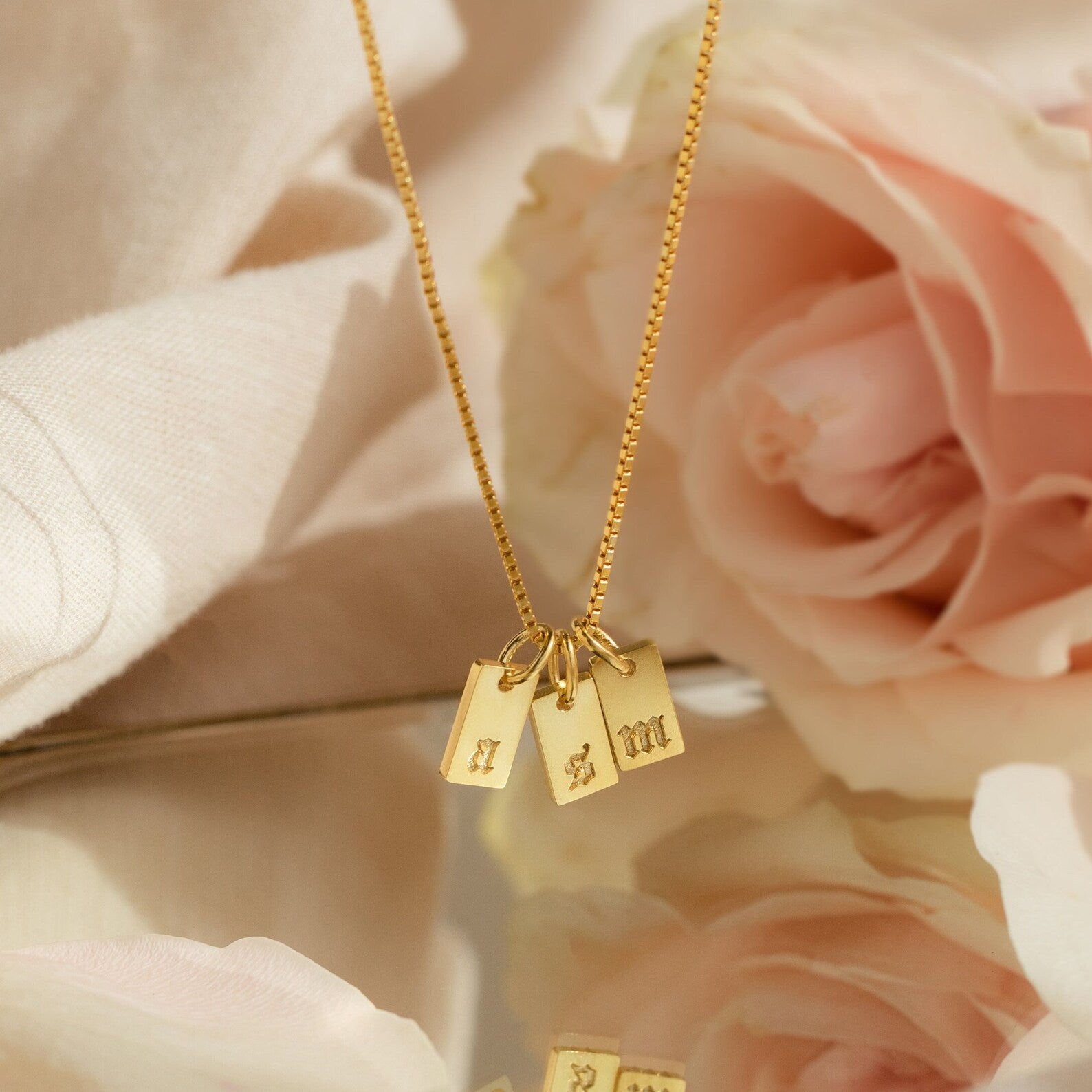 Old English Mini Tag Necklace with three engraved letter charms, elegantly displayed on soft fabric with pink roses in the background for a personalized touch.