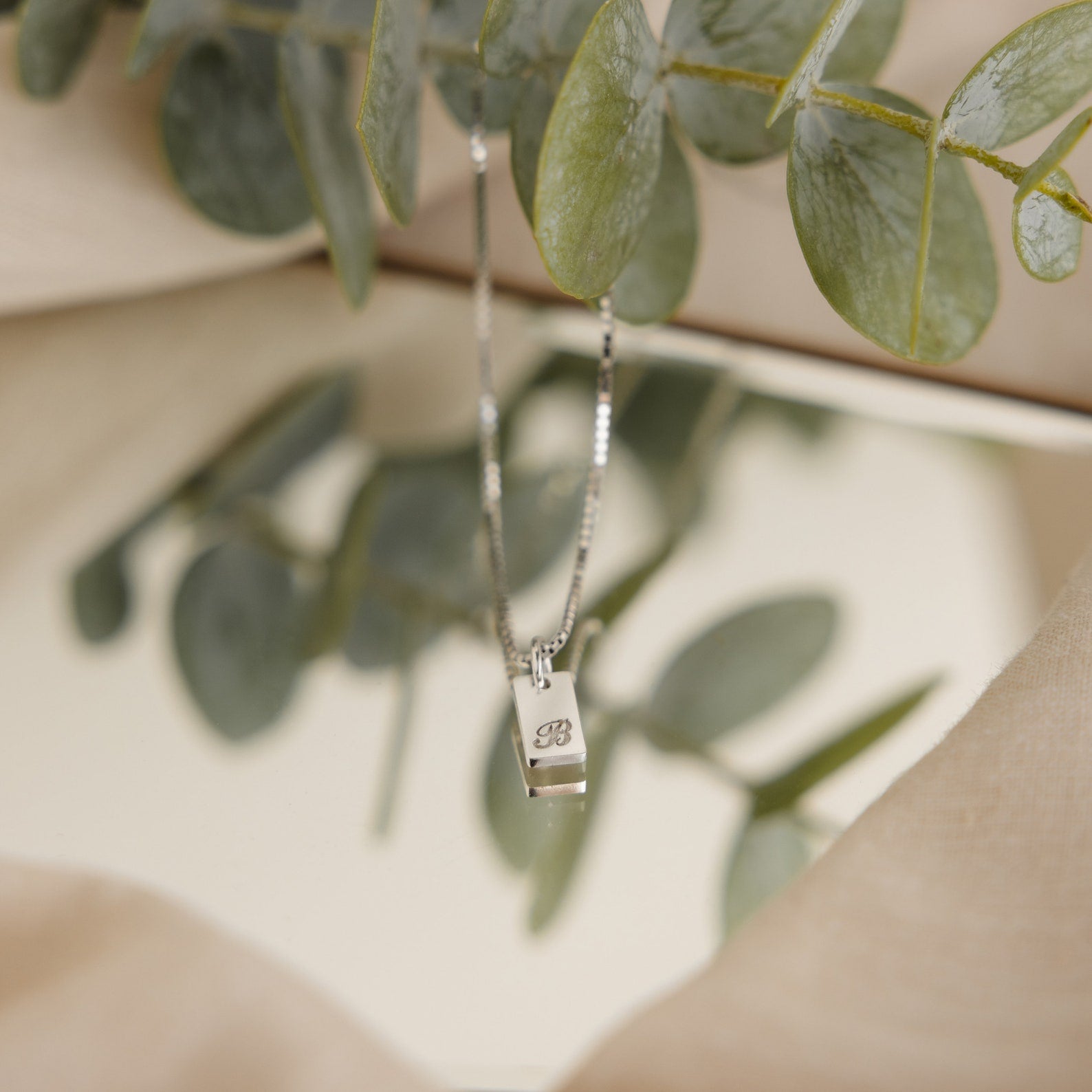 The Carrie Mini Tag Necklace, a minimalist silver initial bar pendant, rests on a mirror surrounded by green eucalyptus leaves—an ideal personalized accessory for a subtle statement.
