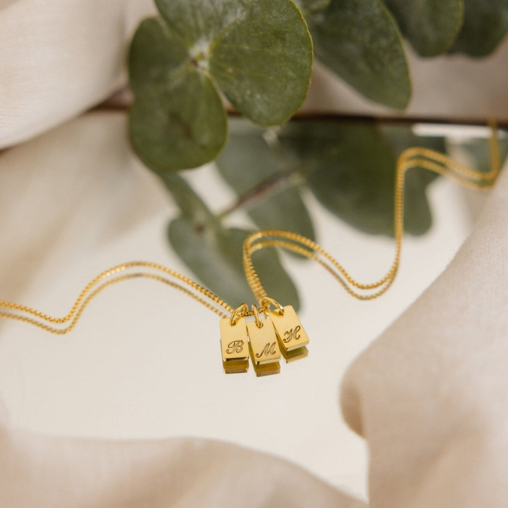 The Carrie Mini Tag Necklace features three engraved rectangular pendants, resting stylishly on a mirror with leaves and fabric in the background—a perfect personalized accessory for any style.