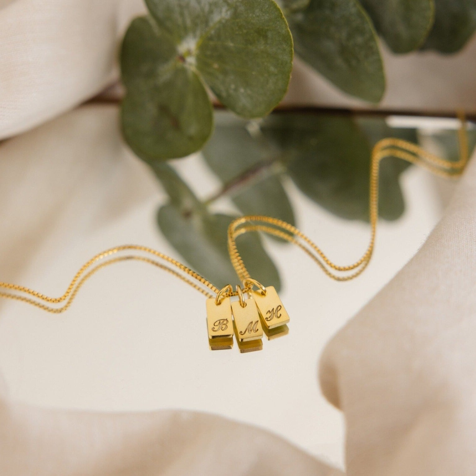 The Carrie Mini Tag Necklace features three engraved rectangular pendants, resting stylishly on a mirror with leaves and fabric in the background—a perfect personalized accessory for any style.