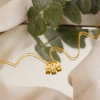 Old English Mini Tag Necklace with three letter charms, shown on a soft surface with green leaves in the background—a stylish personalized necklace for any occasion.