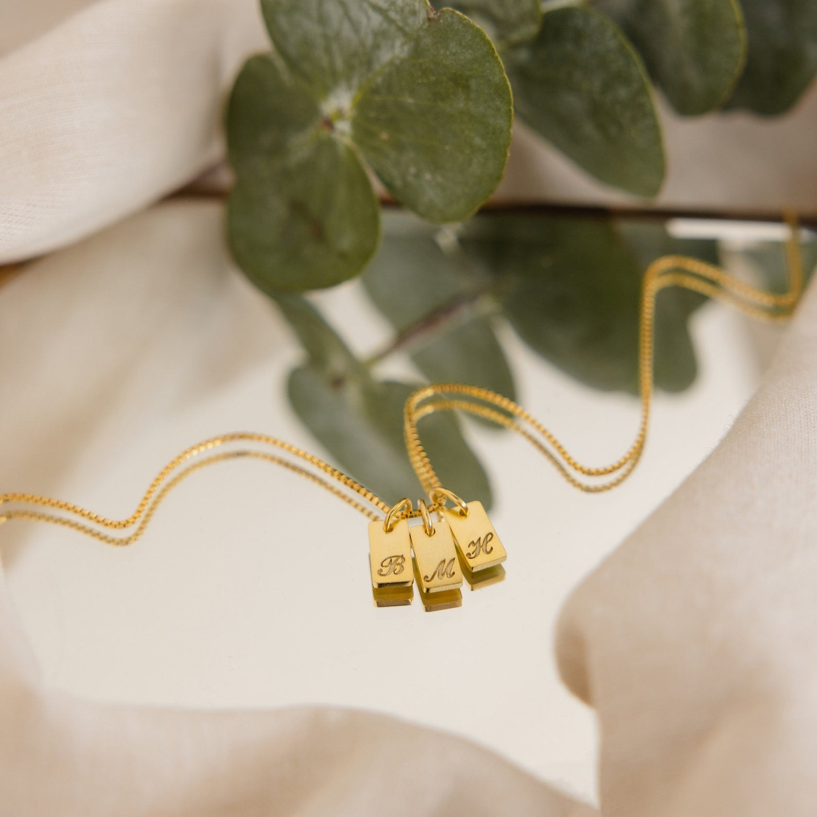 Old English Mini Tag Necklace with three letter charms, shown on a soft surface with green leaves in the background—a stylish personalized necklace for any occasion.