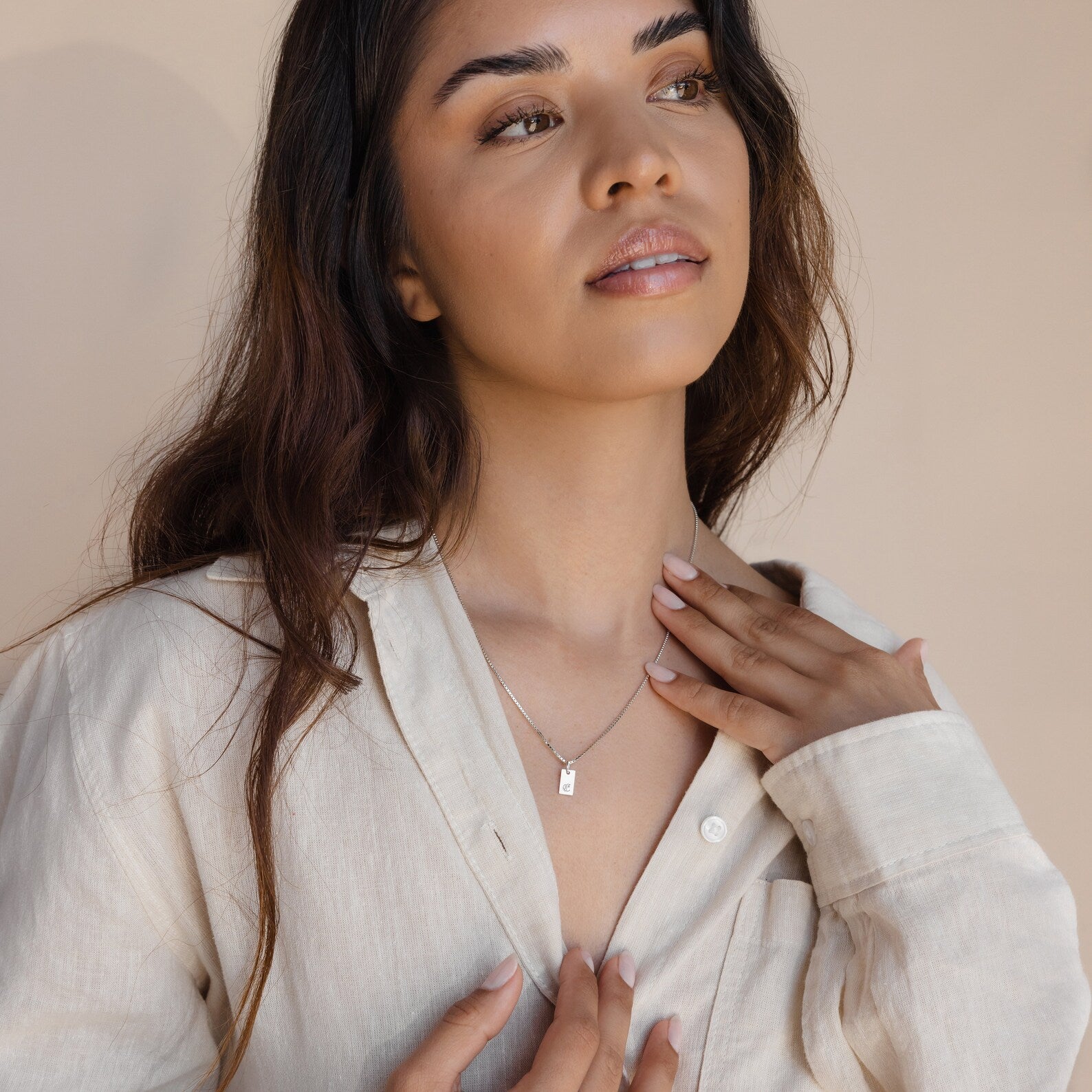 A woman with long brown hair in a beige shirt touches her Carrie Mini Tag Necklace, gazing upward with a soft expression.