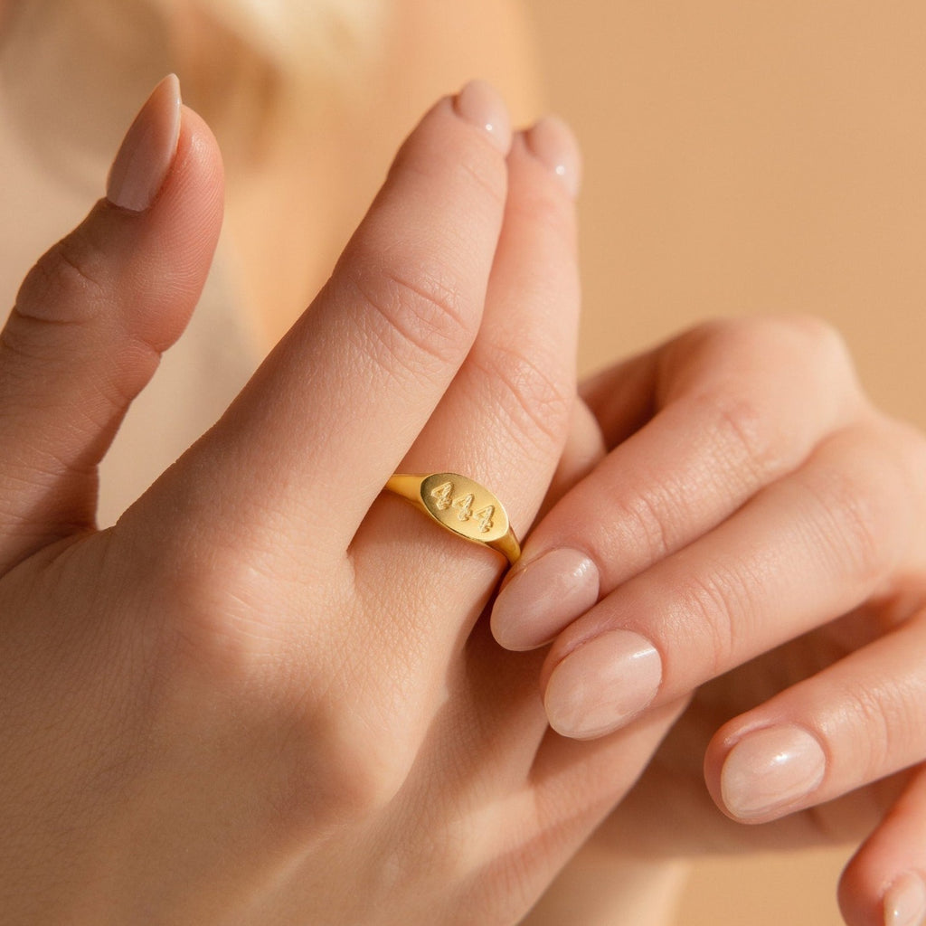 A hand with nude nails wears a minimalist gold Old English Signet Ring engraved with the numbers 444 on the index finger.