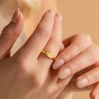 A hand with nude nails wears a minimalist gold Old English Signet Ring engraved with the numbers 444 on the index finger.