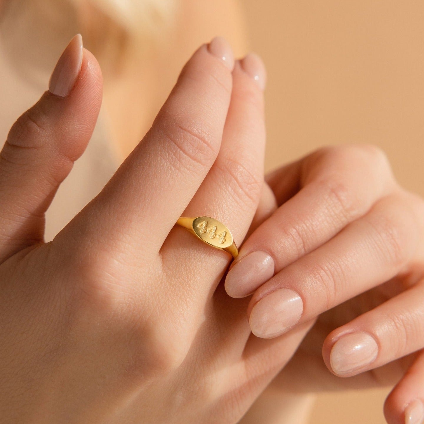 A hand with nude nails wears a minimalist gold Old English Signet Ring engraved with the numbers 444 on the index finger.