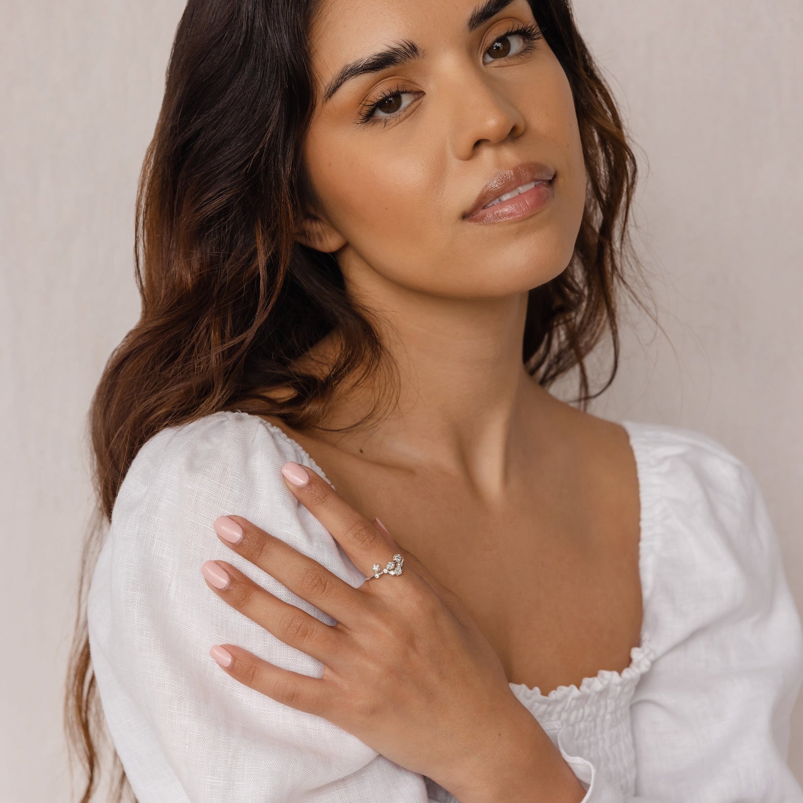 Woman with long, wavy brown hair in a white top showcases the Constellation Ring, posing with her hand near her shoulder.