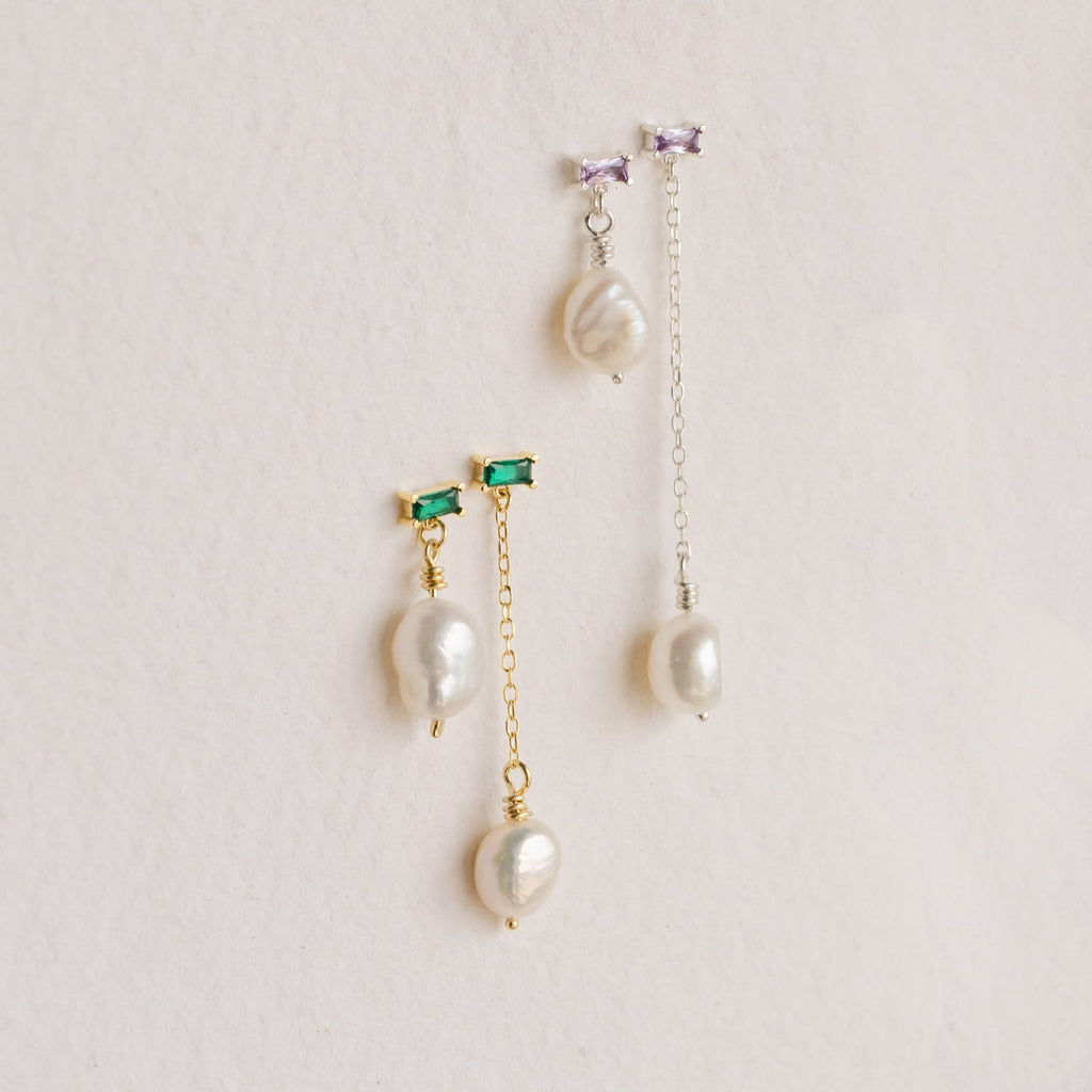 The Asymmetrical Birthstone Drop Earrings feature two mismatched pearl drops—one gold with a green stone, one silver with a purple stone—creating unique custom earrings, displayed on a light background.