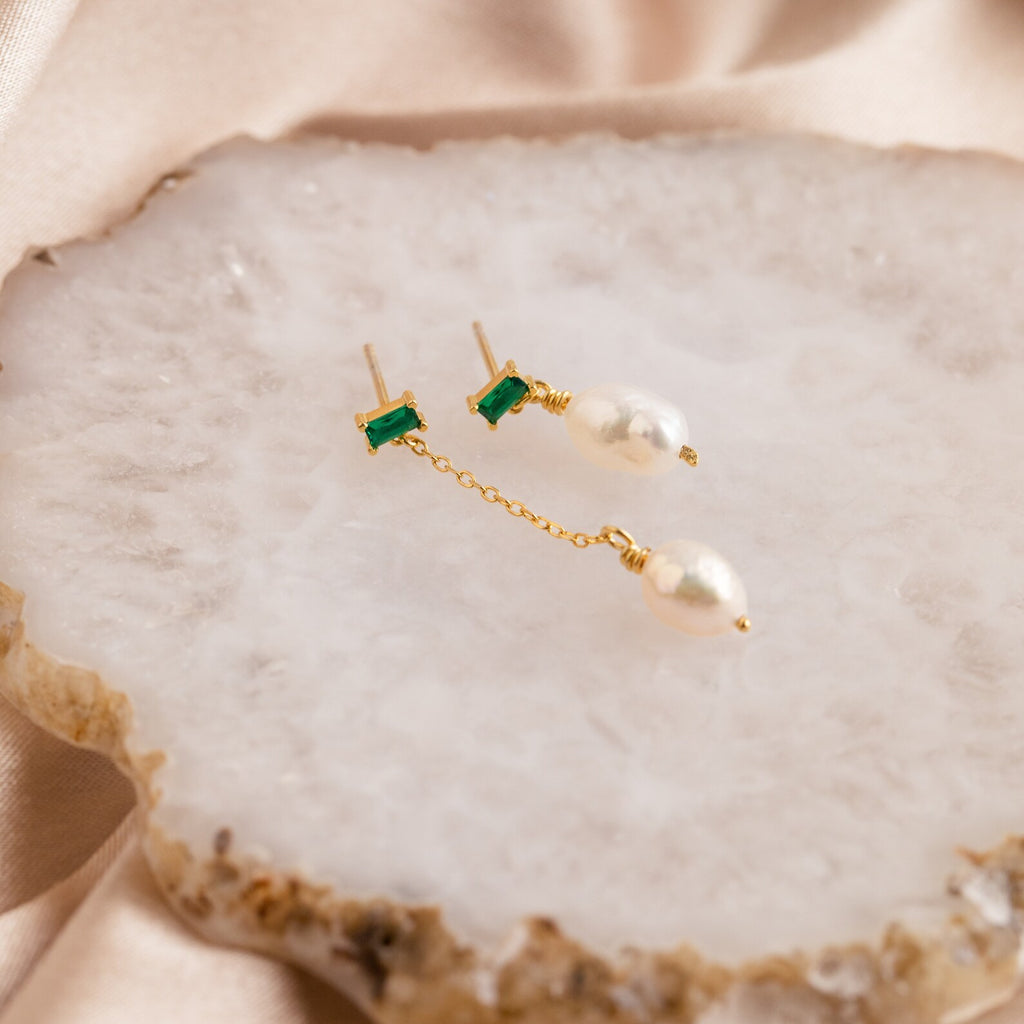 The Asymmetrical Birthstone Drop Earrings feature gold mismatched designs with green stones and dangling pearls, displayed on a white textured stone surface—ideal for custom birthstone jewelry.