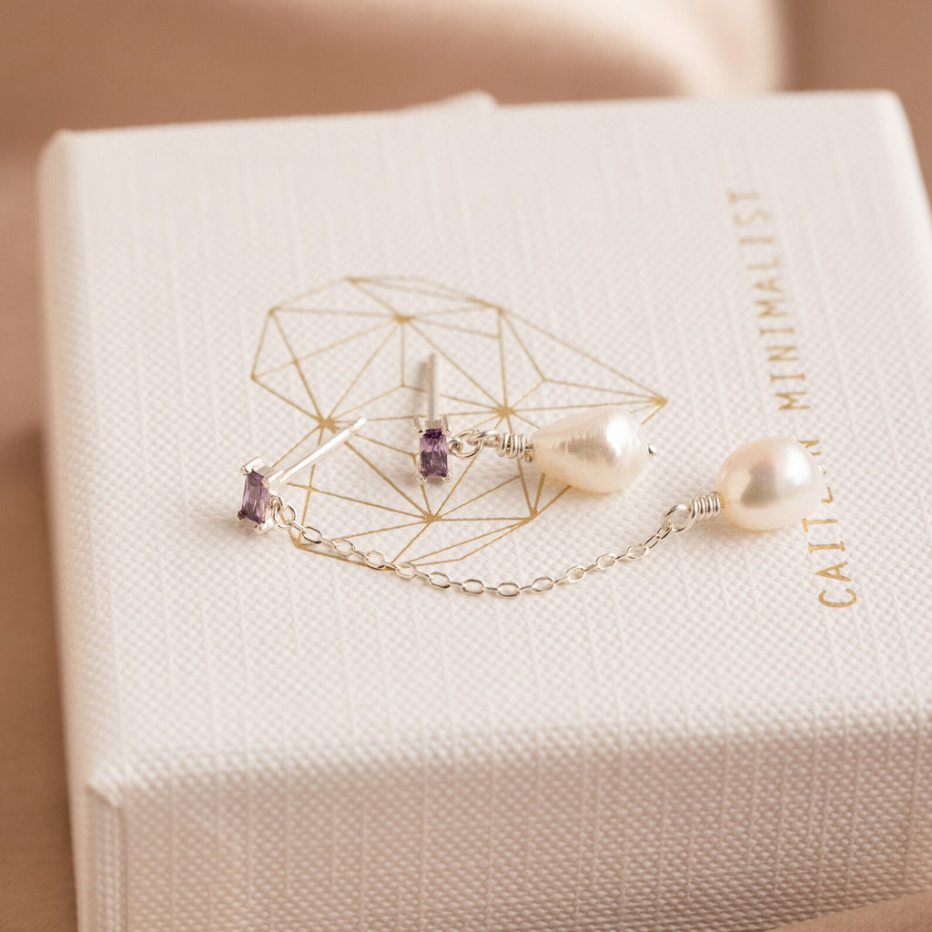 Asymmetrical Birthstone Drop Earrings featuring silver pearl stones and amethyst accents are displayed on a textured white jewelry box with gold details, emphasizing the sophisticated design of these custom earrings.