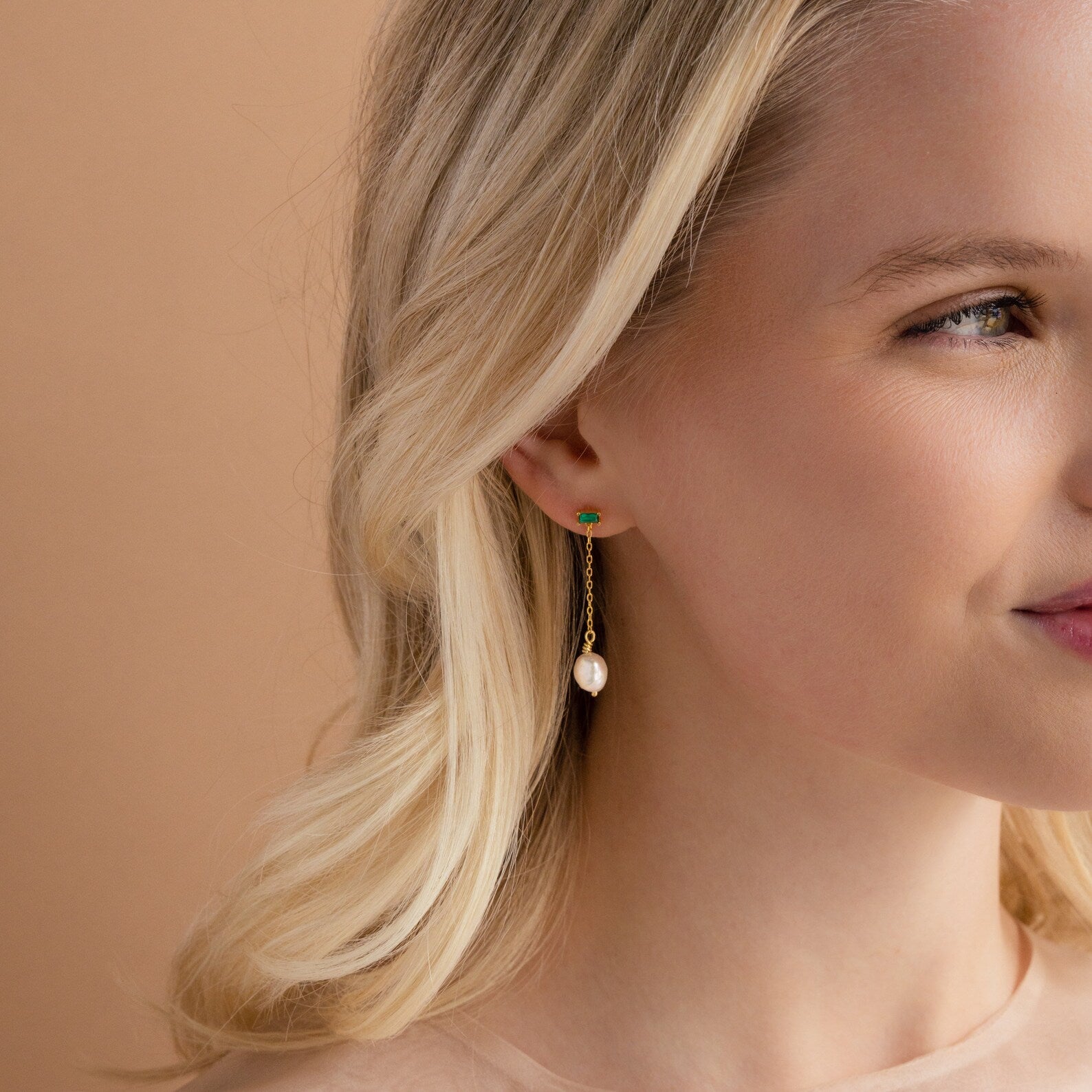 A blonde woman wears Asymmetrical Birthstone Drop Earrings featuring a gold design, green stud, and dangling pearl against a beige background.