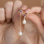 A hand displays Asymmetrical Birthstone Drop Earrings with mismatched pearls, silver chain accents, and small purple stones.