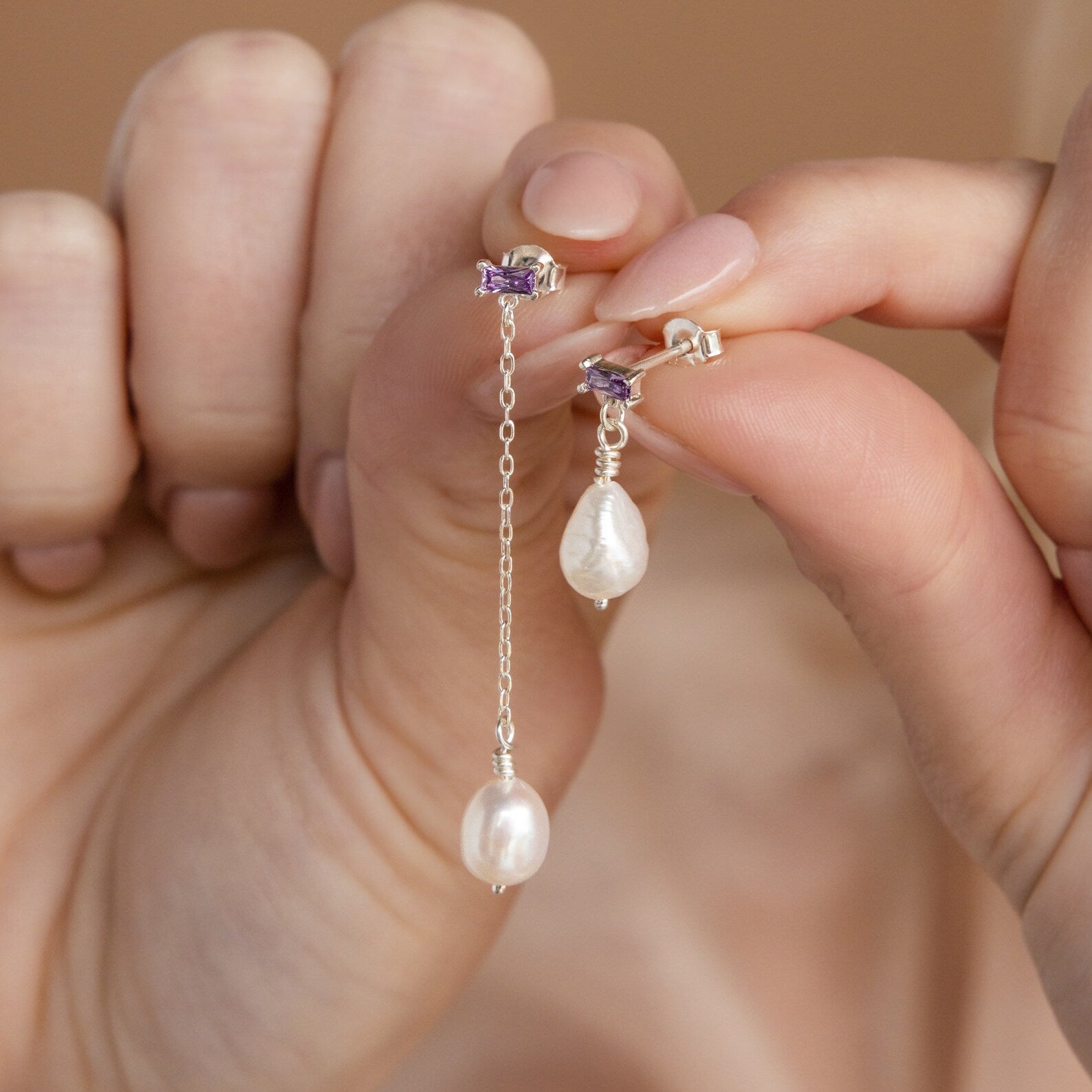 A hand displays Asymmetrical Birthstone Drop Earrings with mismatched pearls, silver chain accents, and small purple stones.