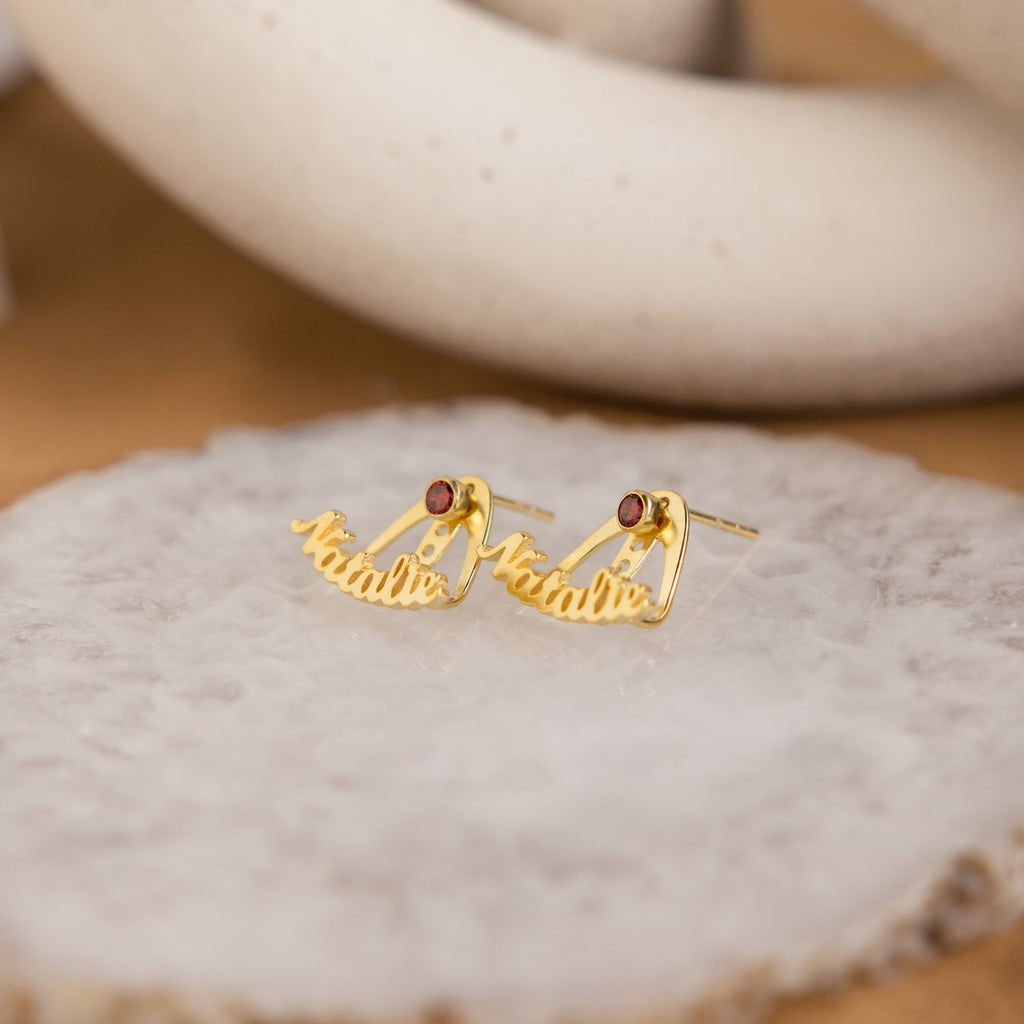London Name Ear Jackets in gold, personalized with "Natalie" and a small red gemstone birthstone, elegantly displayed on marble. These custom birthstone earrings add a unique and meaningful touch to your jewelry collection.