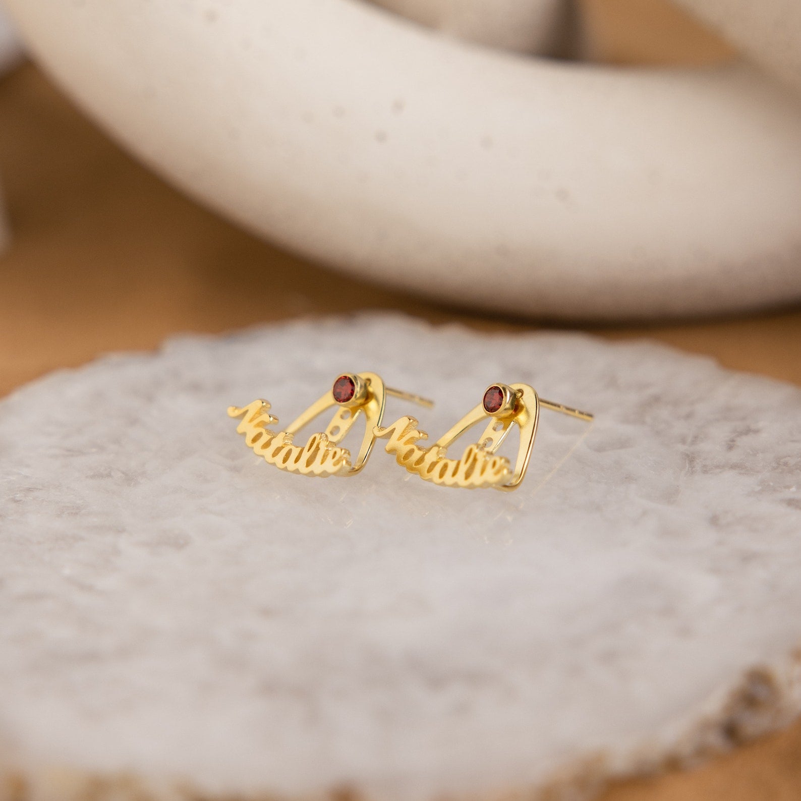 London Name Ear Jackets in gold, personalized with "Natalie" and a small red gemstone birthstone, elegantly displayed on marble. These custom birthstone earrings add a unique and meaningful touch to your jewelry collection.