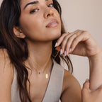 A woman wearing the Newsletter Oval Tag Necklace and a beige top poses with her hand near her chin, looking at the camera.