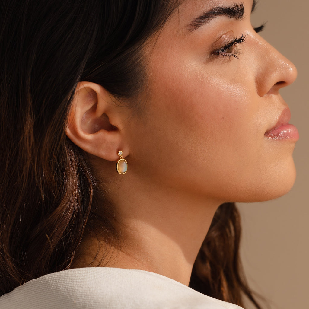A woman in profile wears Moonstone Drop Earrings, featuring a minimalist gold finish, set against a neutral background.
