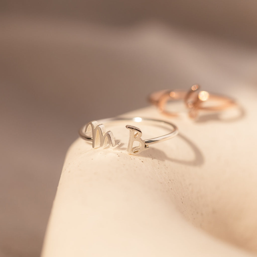 Close-up of the Nella Duo Initial Ring featuring the initials M and B on a soft beige surface—a perfect piece of personalized jewelry.