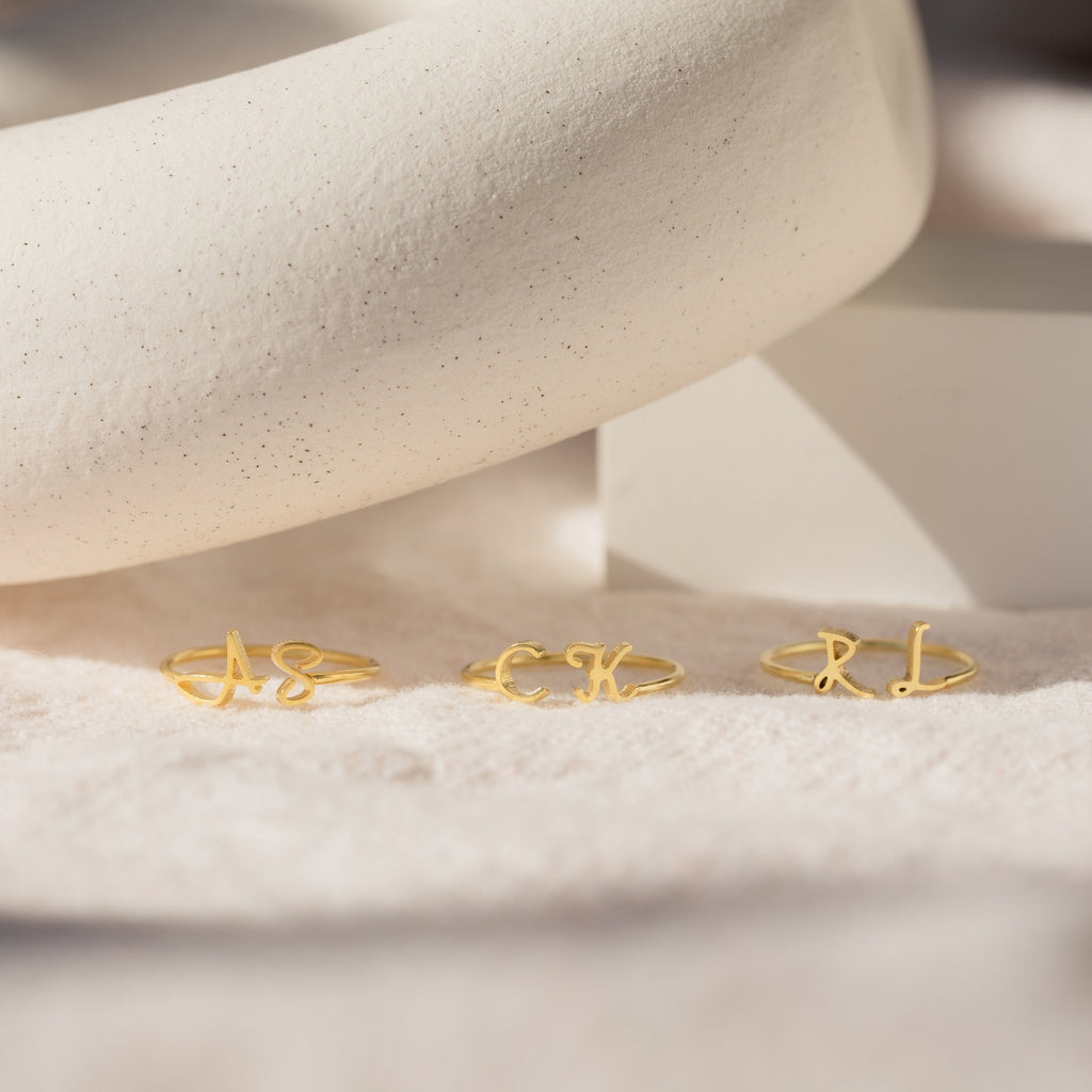 Three Nella Duo Initial Rings with AS, CK, and RL are displayed on a soft beige surface with a white prop—personalized jewelry that's perfect as an anniversary gift.
