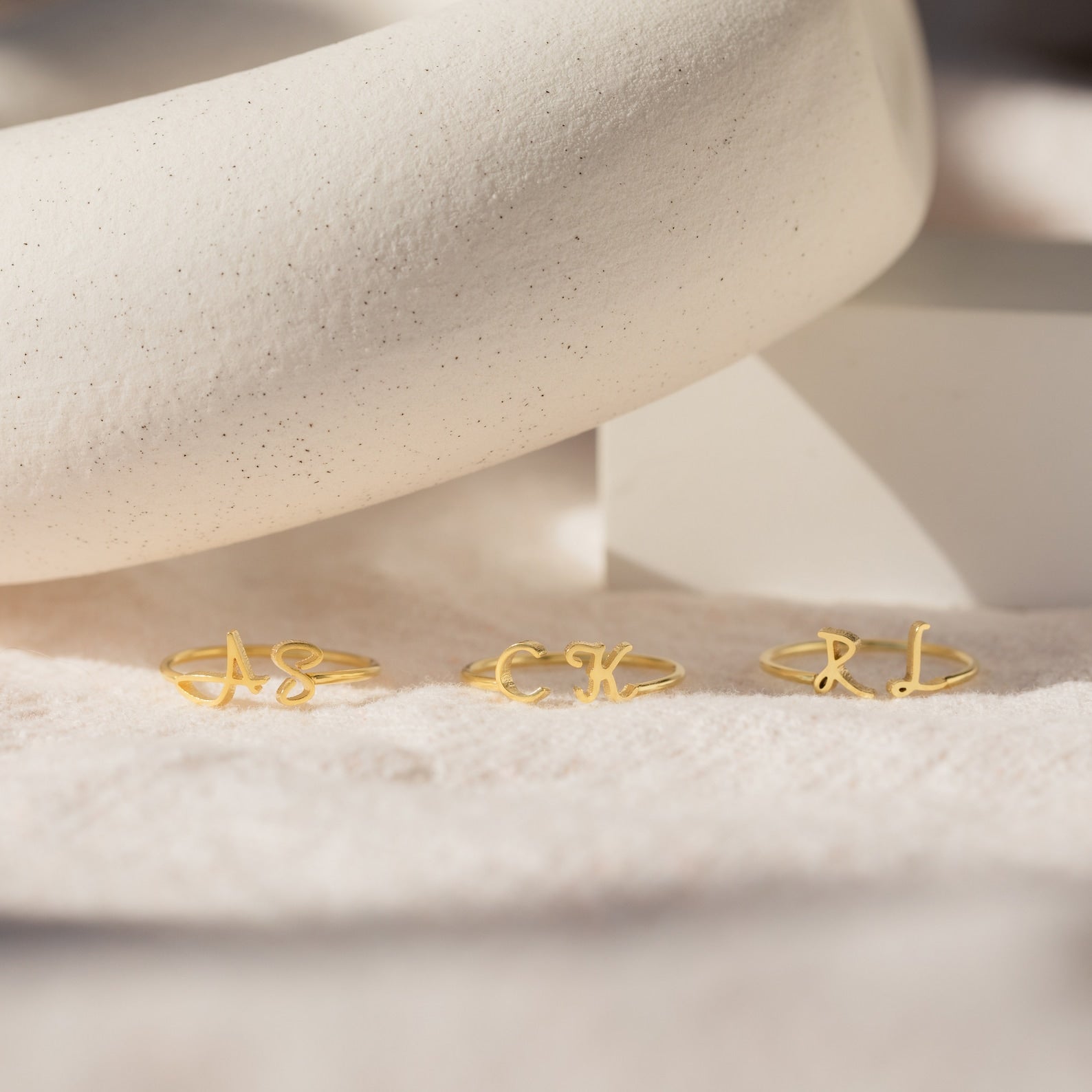 Three Nella Duo Initial Rings with AS, CK, and RL are displayed on a soft beige surface with a white prop—personalized jewelry that's perfect as an anniversary gift.