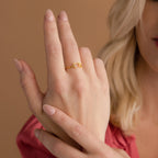 A woman wearing the Nella Duo Initial Ring on her finger, with nude nails and a pink blouse—a stylish, personalized accessory ideal for anniversaries.