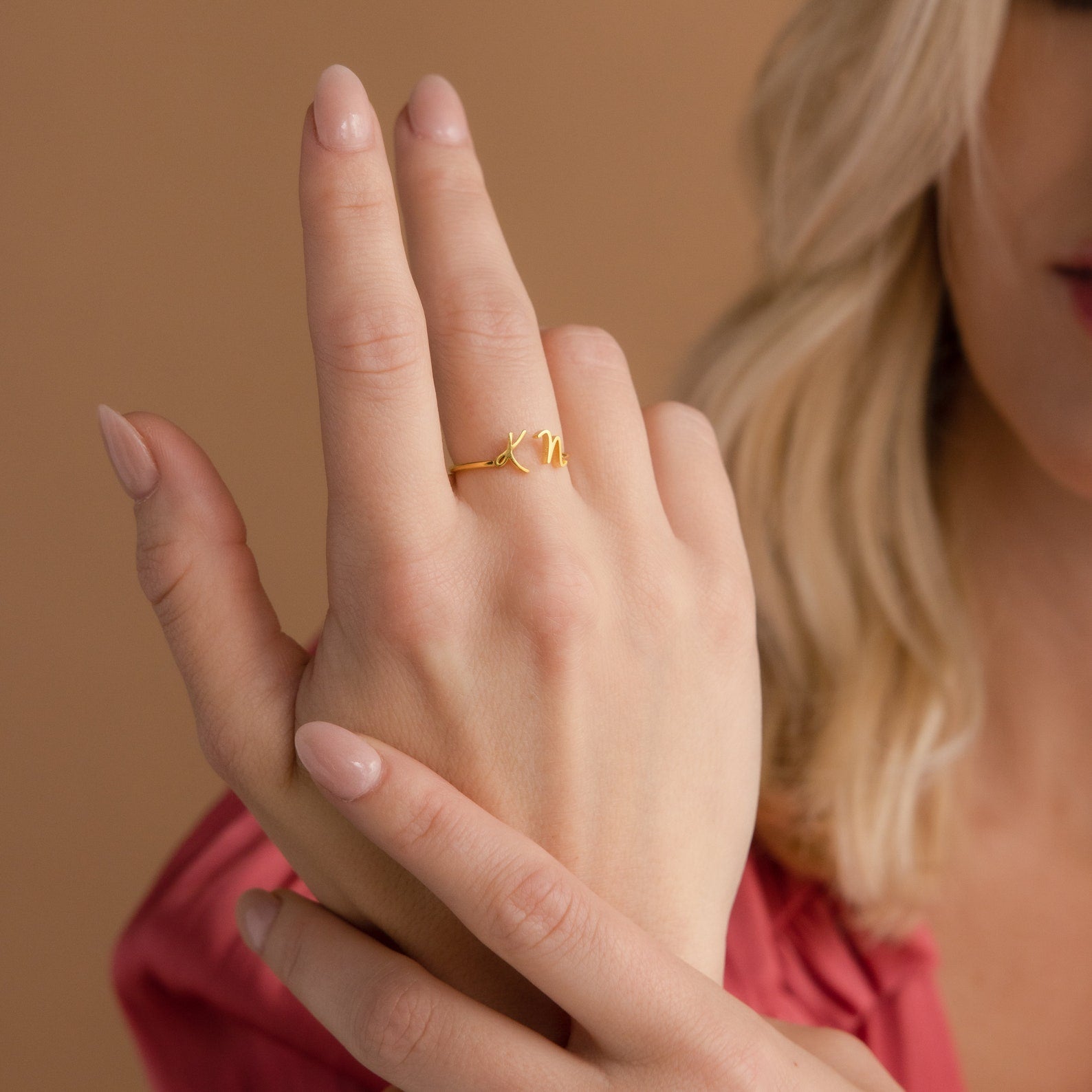 A woman wearing the Nella Duo Initial Ring on her finger, with nude nails and a pink blouse—a stylish, personalized accessory ideal for anniversaries.