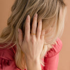 A blonde woman in a pink top covers her face with her hand, highlighting the Nella Duo Initial Ring—a personalized piece perfect as an anniversary gift.