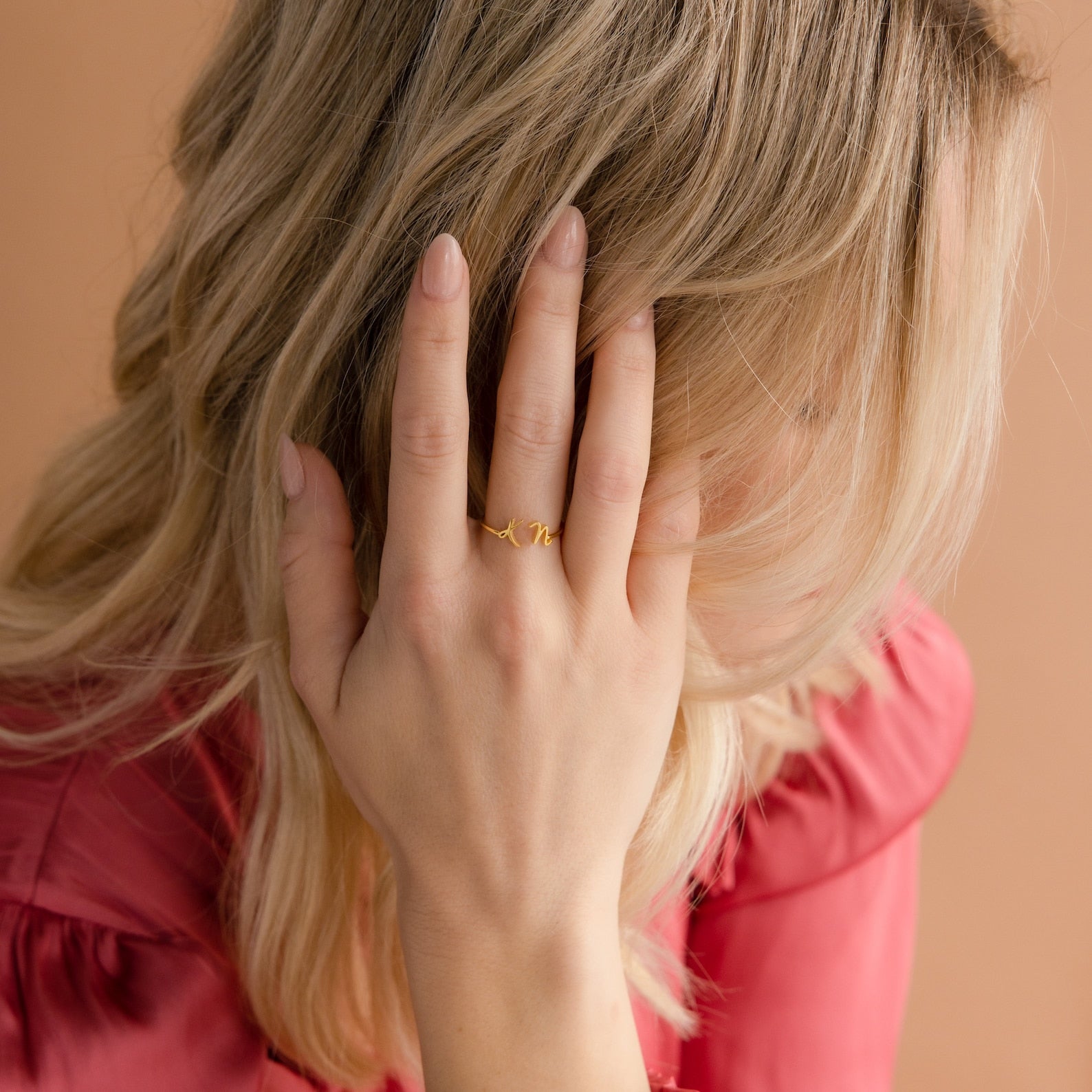 A blonde woman in a pink top covers her face with her hand, highlighting the Nella Duo Initial Ring—a personalized piece perfect as an anniversary gift.