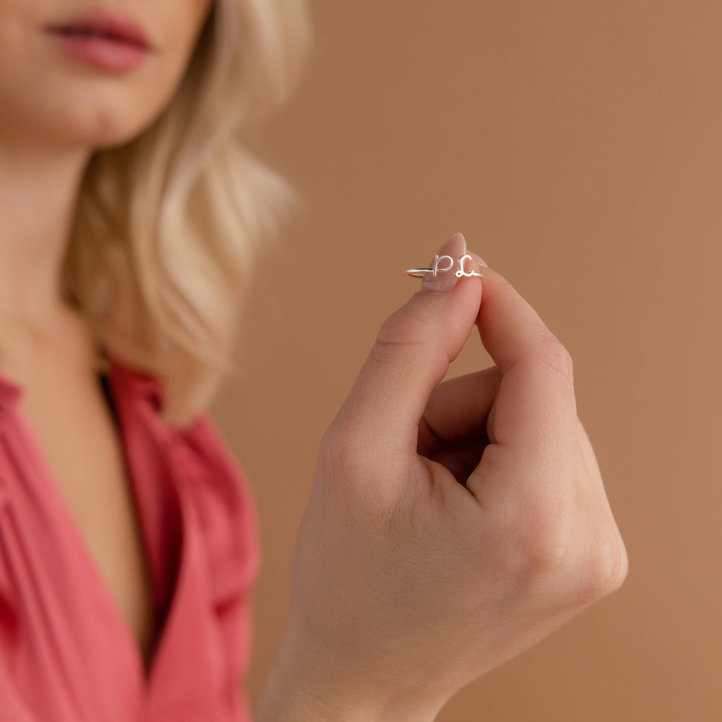 A woman in a pink top holds the Dainty Duo Initial Ring with a script design, making it a perfect anniversary gift, against a beige background.