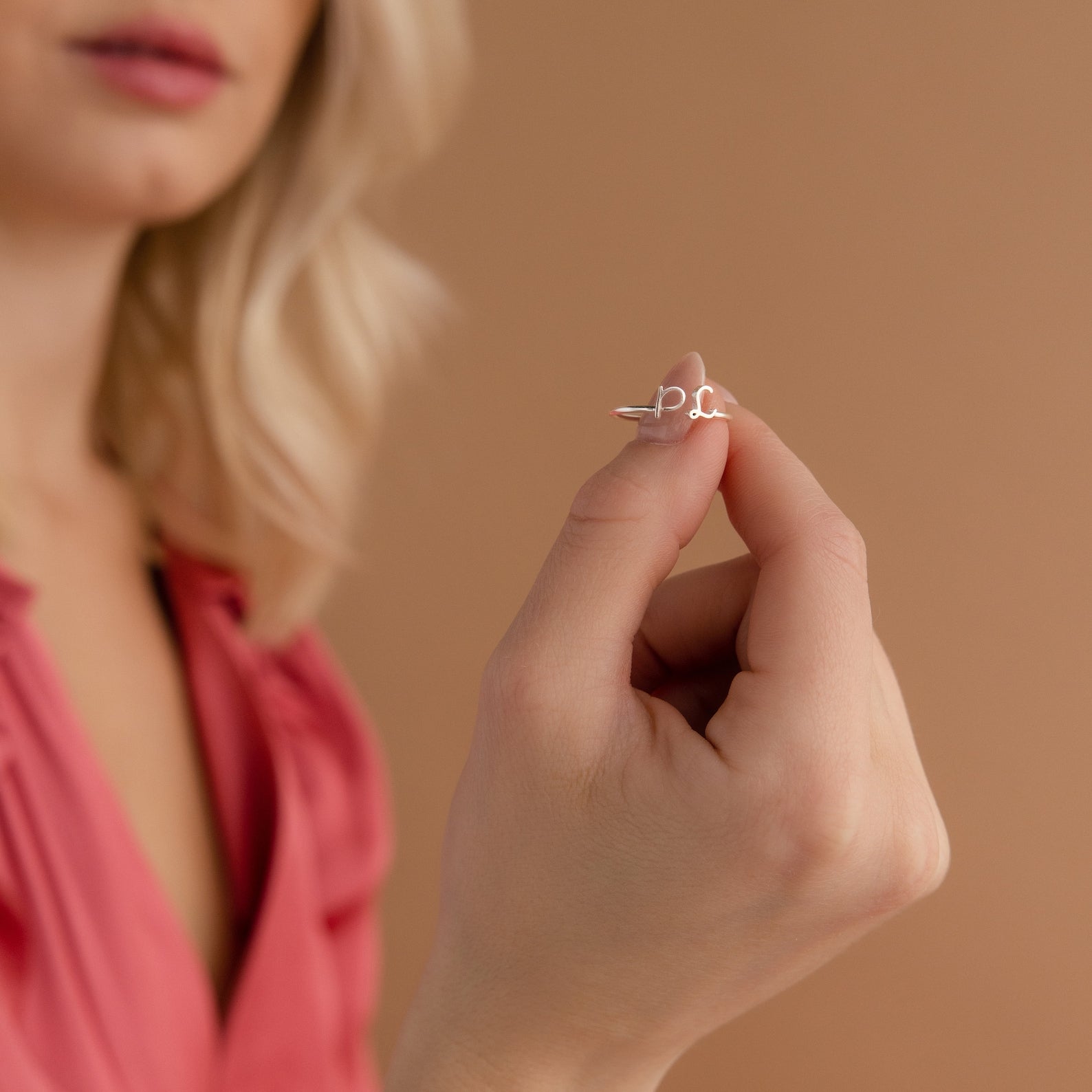 A woman in a pink top holds the Dainty Duo Initial Ring with a script design, making it a perfect anniversary gift, against a beige background.