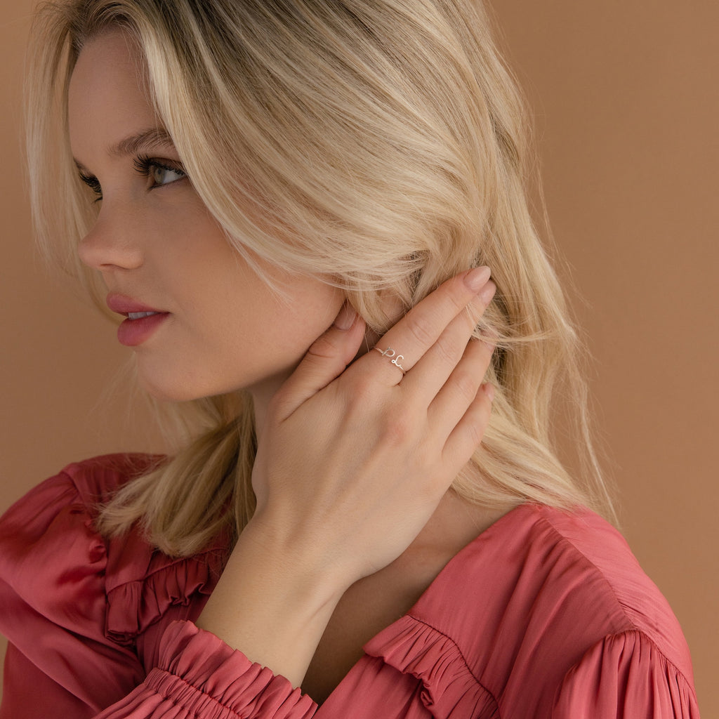 Blonde woman in a pink blouse touches her hair, showcasing the Dainty Duo Initial Ring—a personalized, elegant ring ideal for anniversaries.