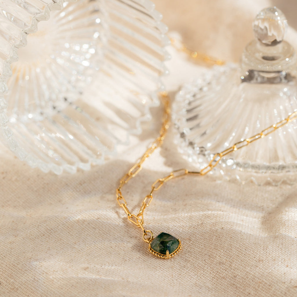 The Agate Pendant Necklace features a gold chain with a green agate pendant, shown beside two clear glass containers on beige fabric—a stunning handmade gemstone accessory.