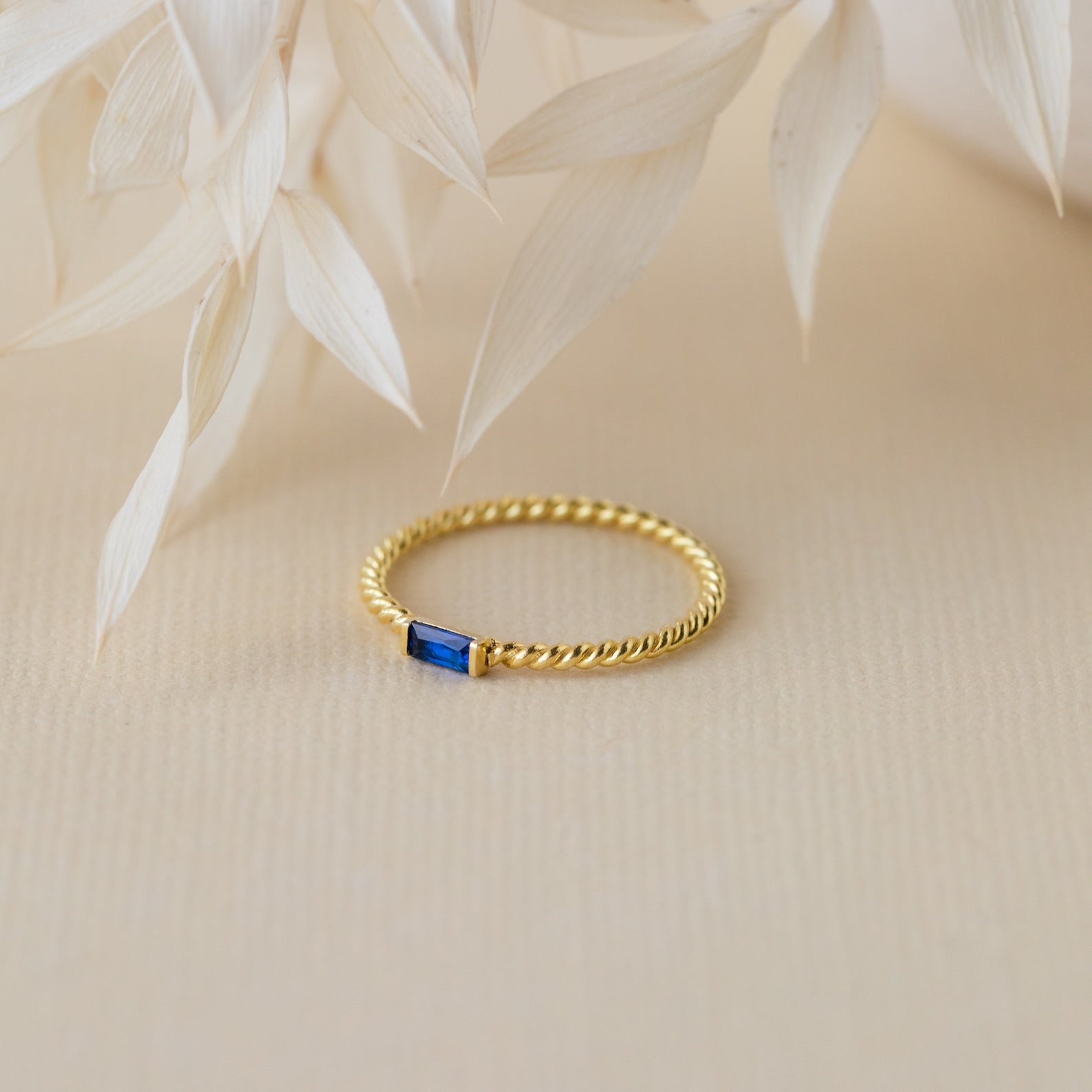 Twist Baguette Birthstone Ring with a small blue rectangular stone, styled on a beige background with dried leaves. Ideal for stacking or adding a personalized birthstone accent to your jewelry collection.