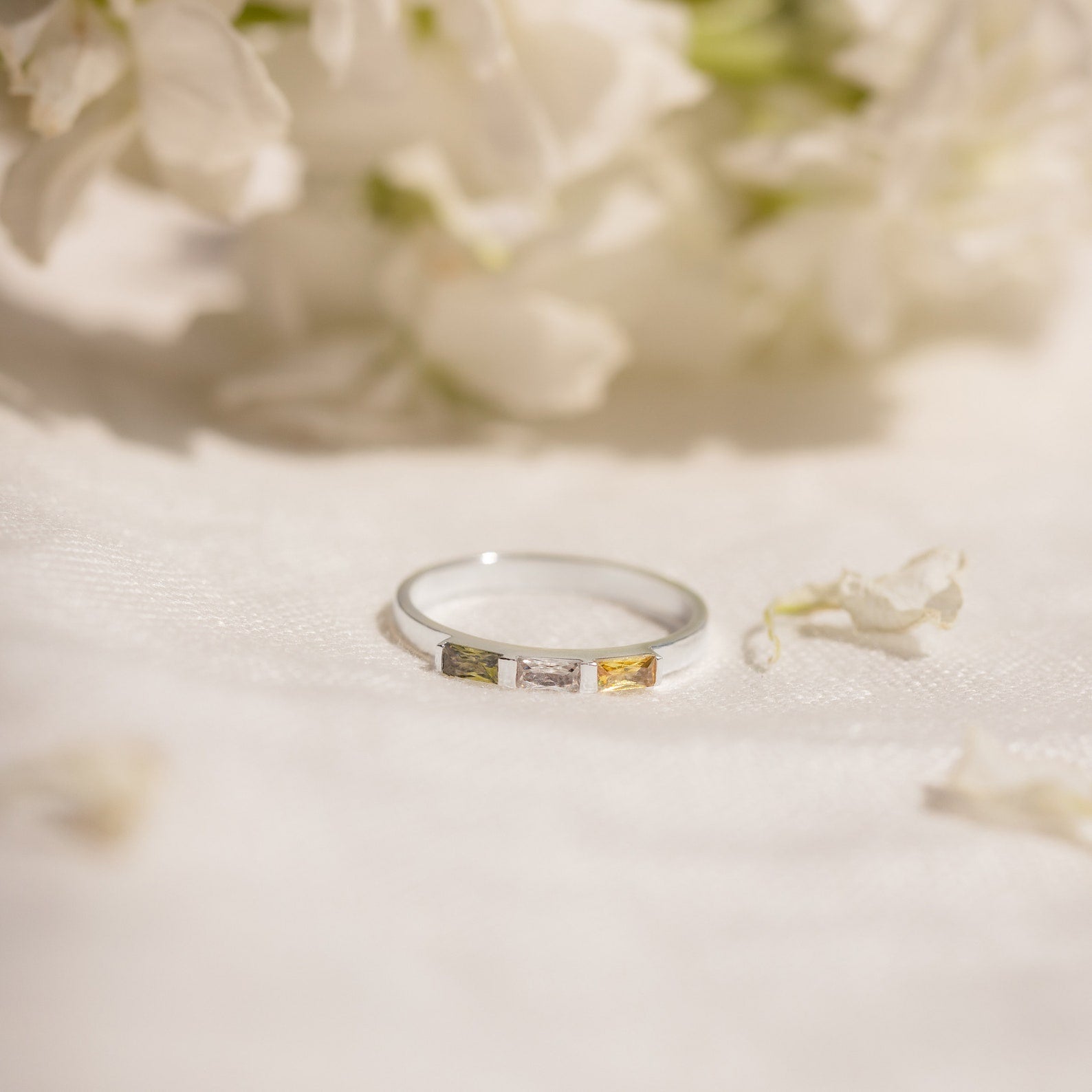 The Mothers Baguette Birthstone Ring, featuring silver and three square stones in yellow, white, and brown, rests on white fabric with a floral backdrop.