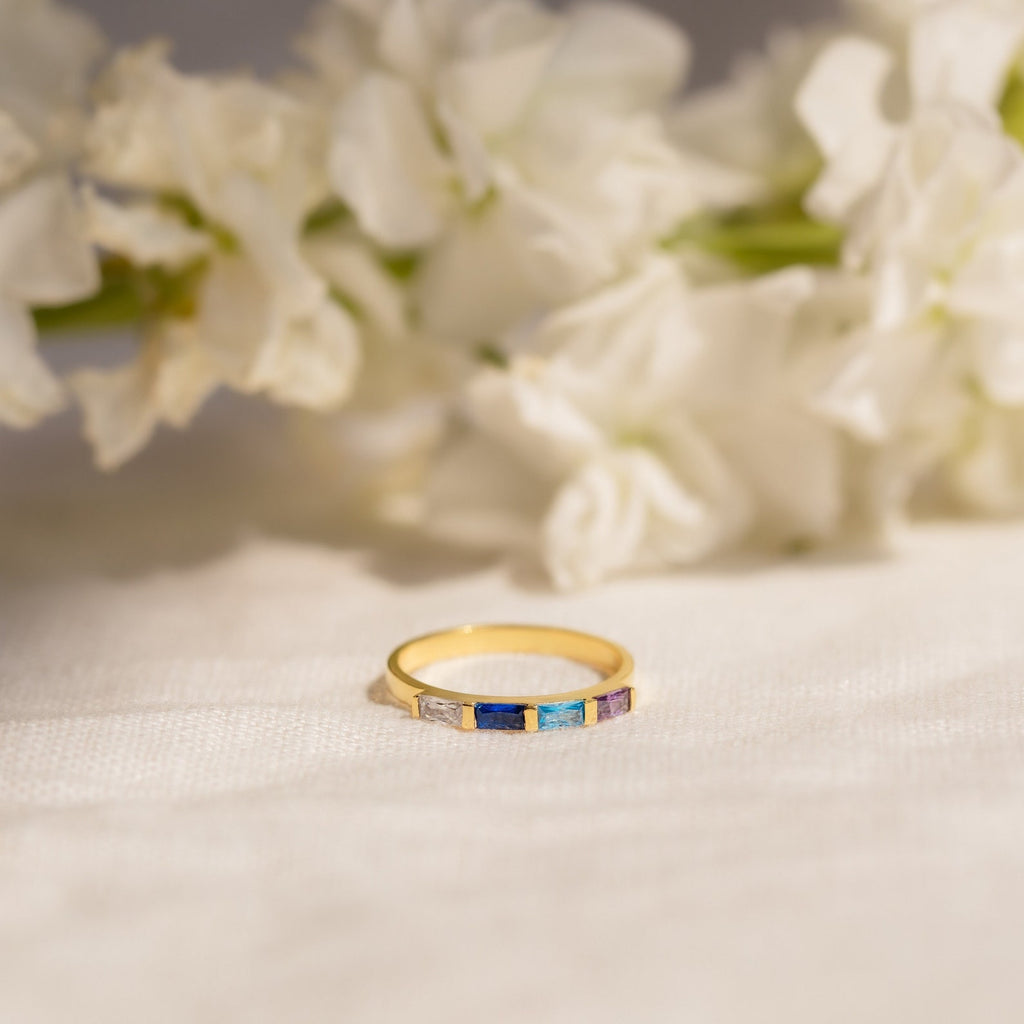 The Mothers Baguette Birthstone Ring, featuring gold with blue and purple stones, is displayed on white fabric with delicate white flowers in the background.