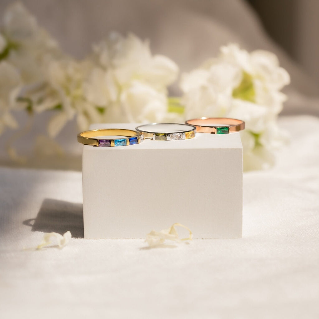 The Mothers Baguette Birthstone Ring, available in gold, silver, and rose gold, is displayed on a white block with delicate white flowers in the background.