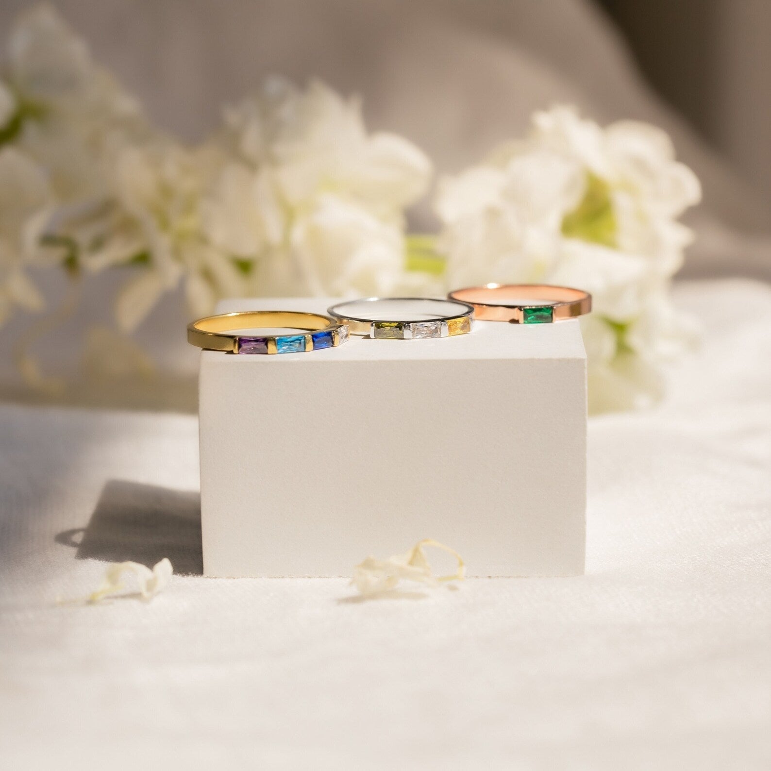 The Mothers Baguette Birthstone Ring, available in gold, silver, and rose gold, is displayed on a white block with delicate white flowers in the background.