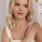 A blonde woman in a white top wears an Initial Morse Birthstone Necklace and a ring, touching her shoulder and smiling softly.