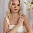 A blonde woman in a white top touches her shoulder, wearing a gold necklace and the Initial Morse Code Ring, with white flowers nearby.