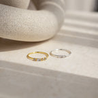 A gold ring and an Initial Morse Code Ring with sparkling stones are displayed on a beige stone surface in natural light, highlighting the elegance of personalized jewelry.