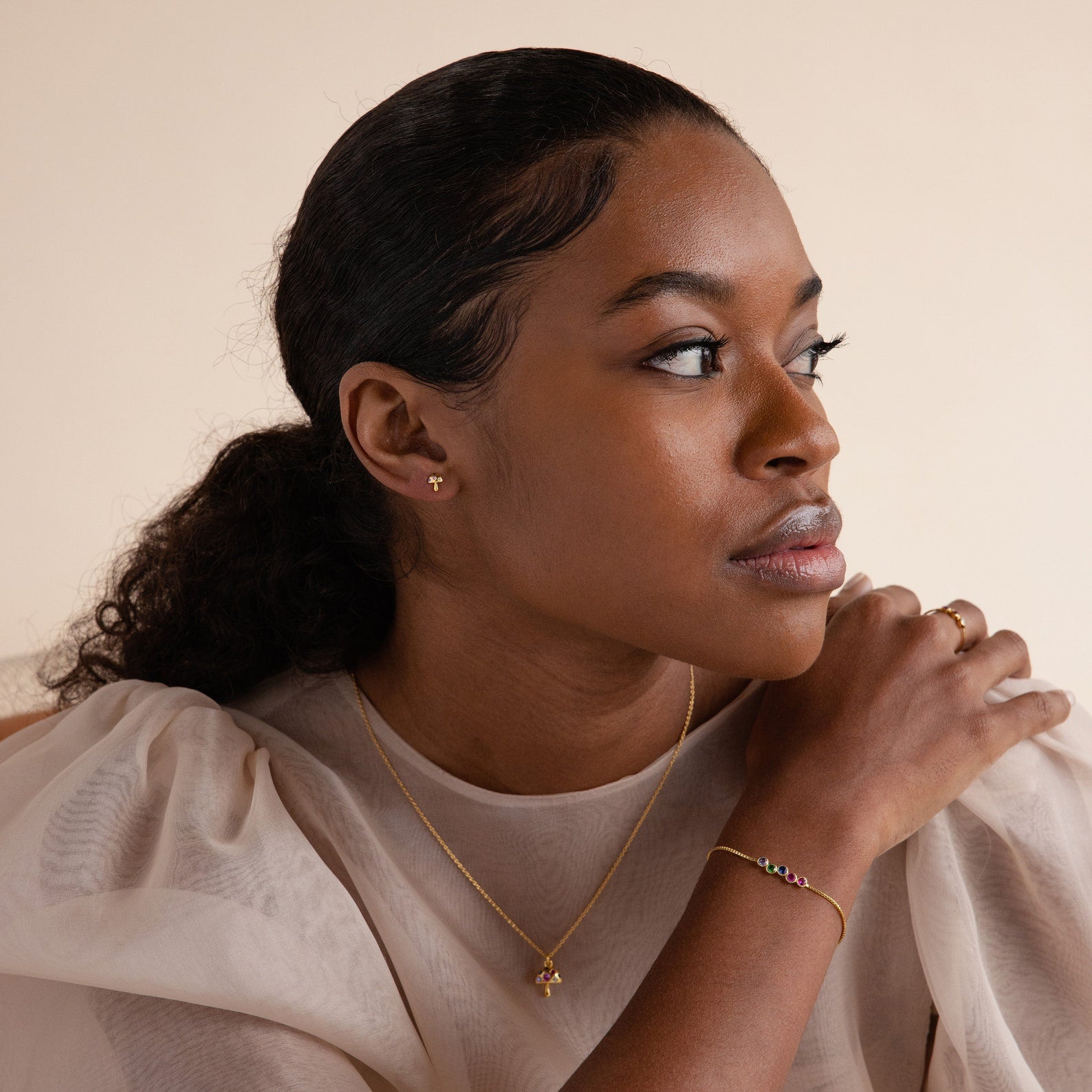 Woman in a cream blouse wears gold jewelry, including Mushroom Studs—inspired by nature—as she gazes to the side with her hand on her shoulder.
