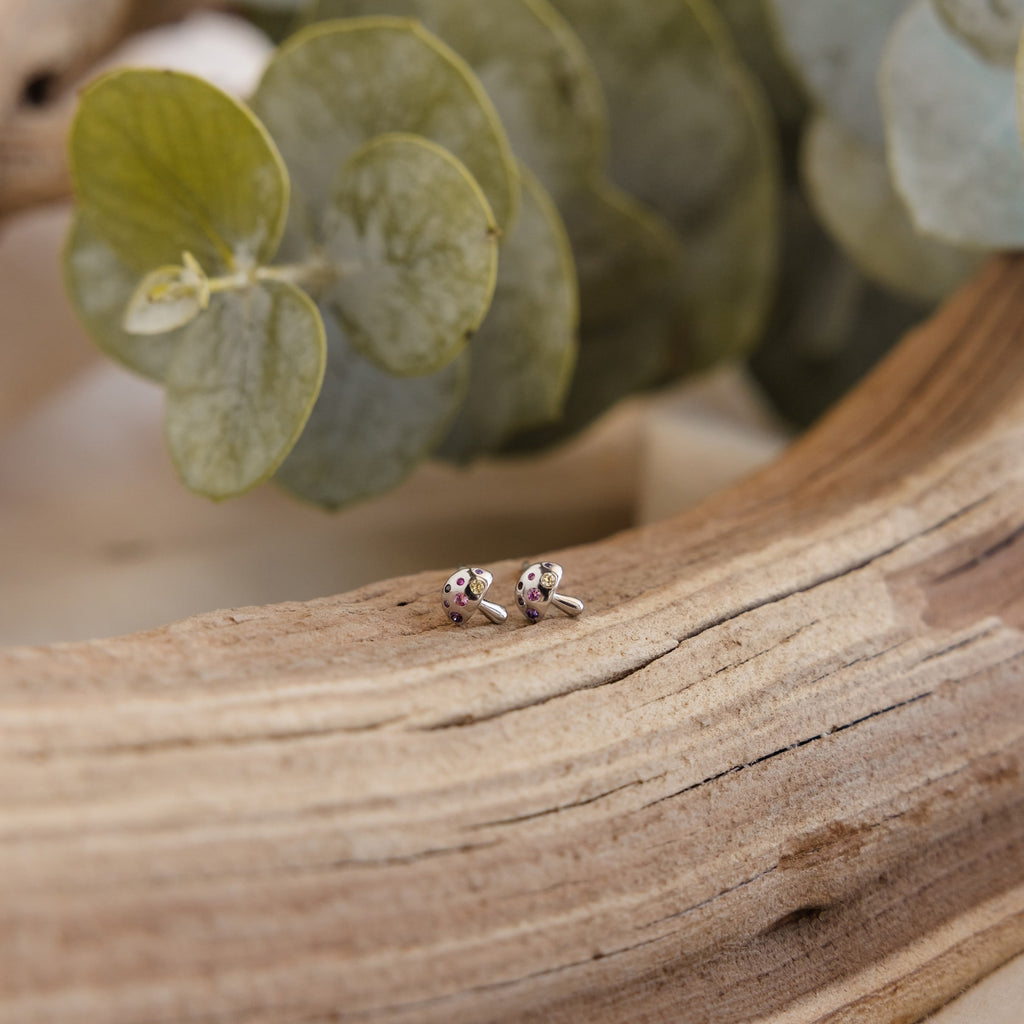 Delicate silver Mushroom Studs with sparkling CZ diamonds rest on a textured wood surface, green leaves in the background—a charming piece of nature-inspired jewelry.